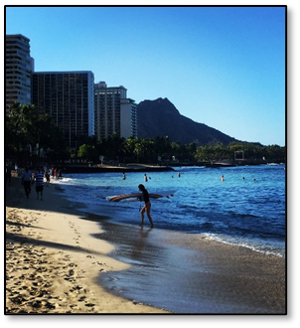 A beach with sand on the left, ocean on the right, a surfer emerging from the water in the midground, and swimmers and buildings in the background