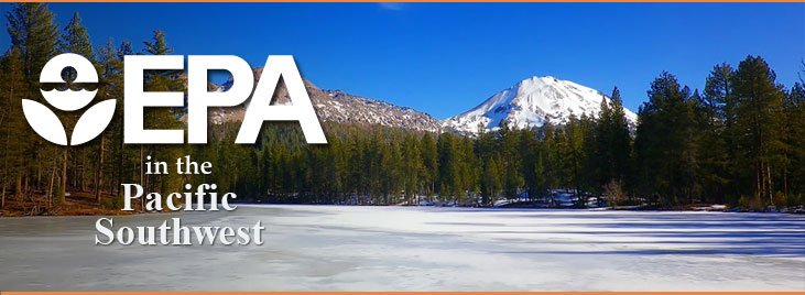 Foreground ice covered lake lined with pine trees with snow covered mountains in background.