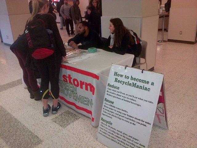 This is a picture of an informational table. Two people are sitting down behind the table, talking to two people standing up in front of it. There's a sign next to the table, which is covered in white cloth, that says: How to become a RecycleManiac.