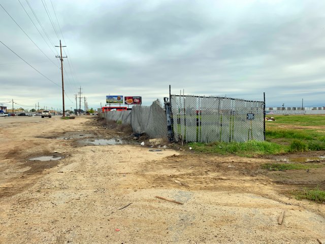 empty lot alongside a road with puddles, chainlink fence and overgrown weeds