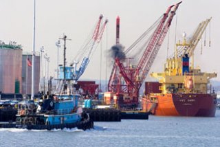 A photograph of a ship, crane, and tugboat with black smoke being emitted.