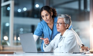 Two women in lab coats sitting in front of laptop screen.