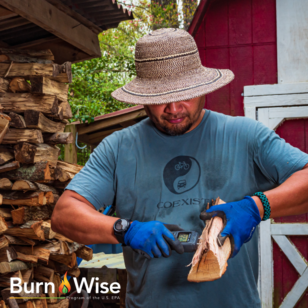 Man standing in front of a woodpile with a piece of firewood in one hand and a moisture meter in the other that is inserted into the firewood to test it.