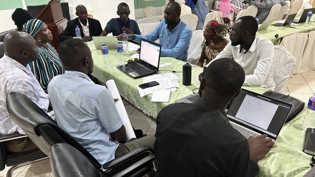 8 men sitting around a table with 2 laptops open and having a group discussion.