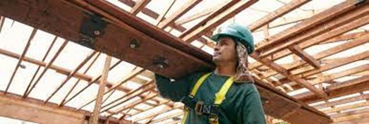 This is a picture of a man with a hard hat on carrying a plank of wood on his shoulder.