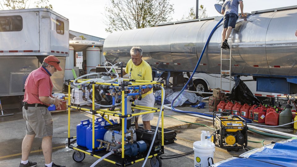 Two men attend to the WOW Cart, a shopping-cart sized cart on wheels. A third person is on a ladder of a tanker truck behind them. 