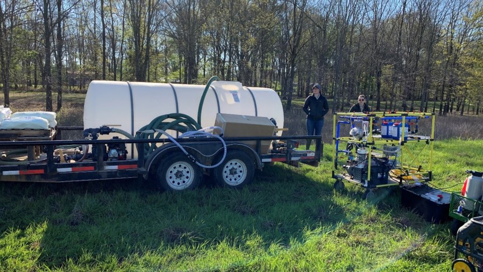 A large white water tank sits on a trailer in a field, with a treeline behind it. The WOW Cart sits to the right of the trailer.