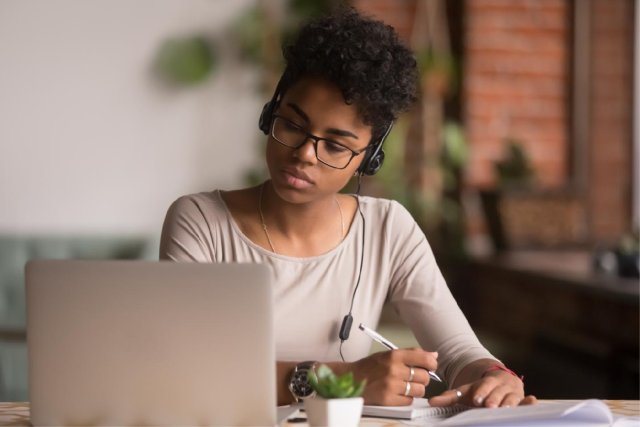 Woman sitting in front of her computer taking notes on a pad of paper while wearing headphones.