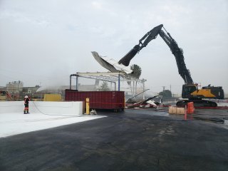 Construction equipment removing corrugated metal roof from building while worker sprays water