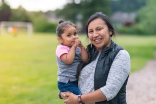 A young woman holds her toddler daughter beside a soccer field.