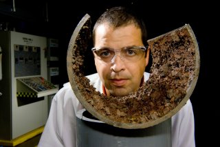 Researcher in a lab coat and wearing safety glasses looks through a pipe covered with corrosion. 