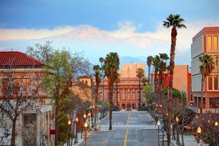 A view of clay colored buildings along a street in San Bernardino with palm trees and a blue sky and white clouds in the background