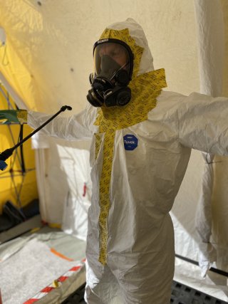 Responder in a Tyvek suit and mask stands in the decontamination line tent with arms stretched waiting to be sprayed with decontaminant.