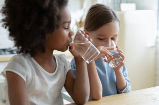 Two children drinking water from glasses while sitting next to each otherat a counter.