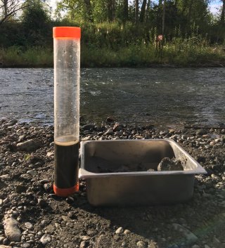 Beaker filled with soil and water standing next to a collection tin. Both are sitting on the bank of a stream.