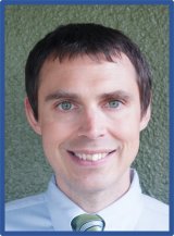 A closeup of a smiling man with short brown hair. He is wearing a collared shirt and tie.