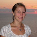 A light-skinned woman with her hair in a low ponytail smiles. An orange sky and sunset is behind her.